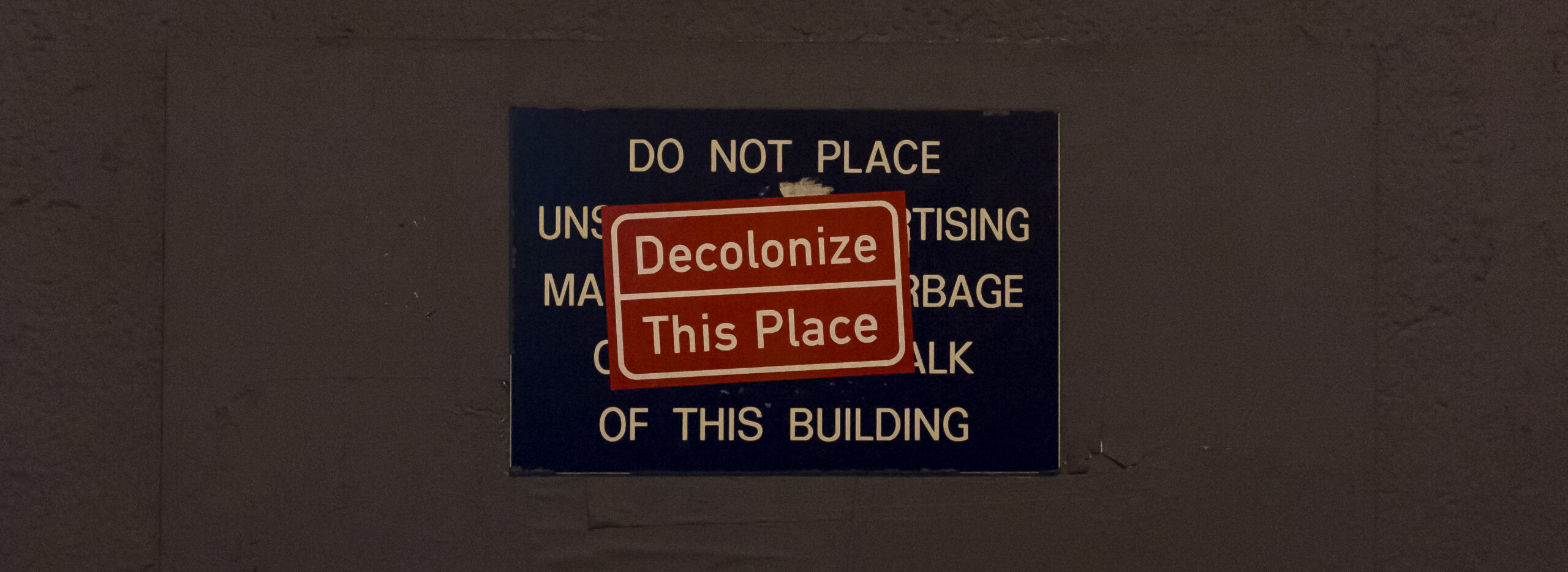A sign on a plain brown wall that reads “do not place...” is obscured by a red sticker saying “Decolonise this place”
